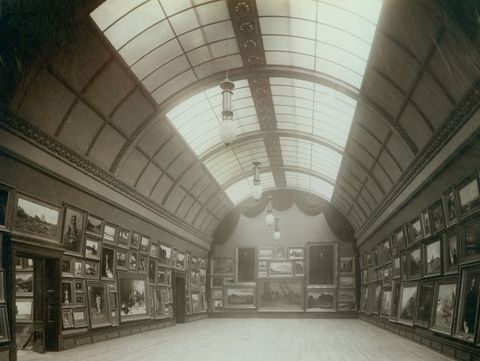 Installation photograph. Black and white photograph shows a long room with an arched, tunnel-like ceiling lined with skylights. Paintings are hung frame-to-frame, three or four levels high. The show history paintings, landscapes, and portraits.