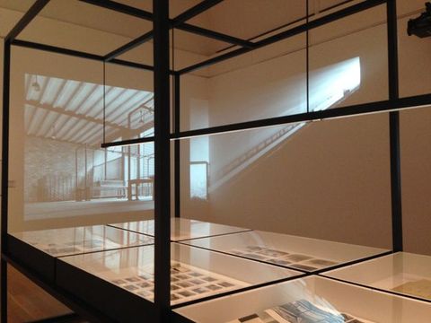Installation photograph. Black and white film projects over glass cases holding printed material and perhaps photographs in the corner of a gallery. One film still shows an empty room. The other looks like the projector is so close to the wall that the image is distorted, becoming a ray of light along one wall and a sliver of film on the other.