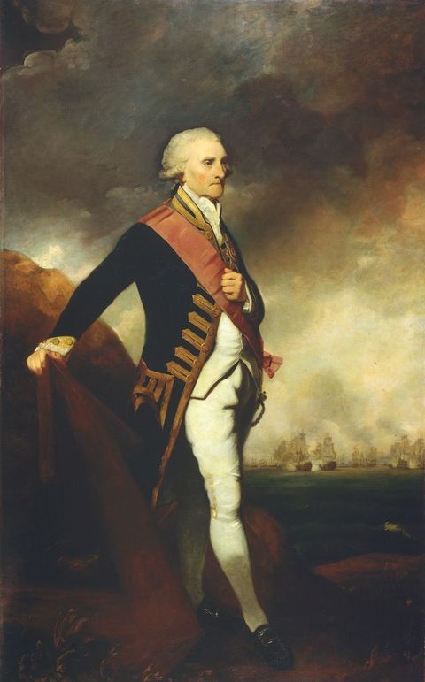 Painting. Full length portrait of a pale-skinned man wearing a military uniform and a white wig stands looking off to our right in front of a seascape under a dark, cloudy sky. One hand rests on the point of an anchor lying in the grass next to him, and the other is held to a dark pink sash that crosses his torso. Masted ships are clustered on the water in the deep distance to our right.