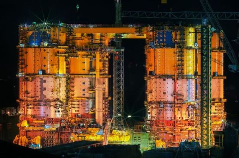 Colour photograph. The attached silo-like structures of an offshore plant are vivid orange, yellow, and blue against a velvet-black sky.