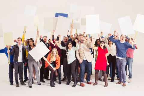 Colour photograph. A group of men and women stand in a tight-knit group and hold up picket signs, all of which are empty. Some raise fists and all shout, mouths wide open.